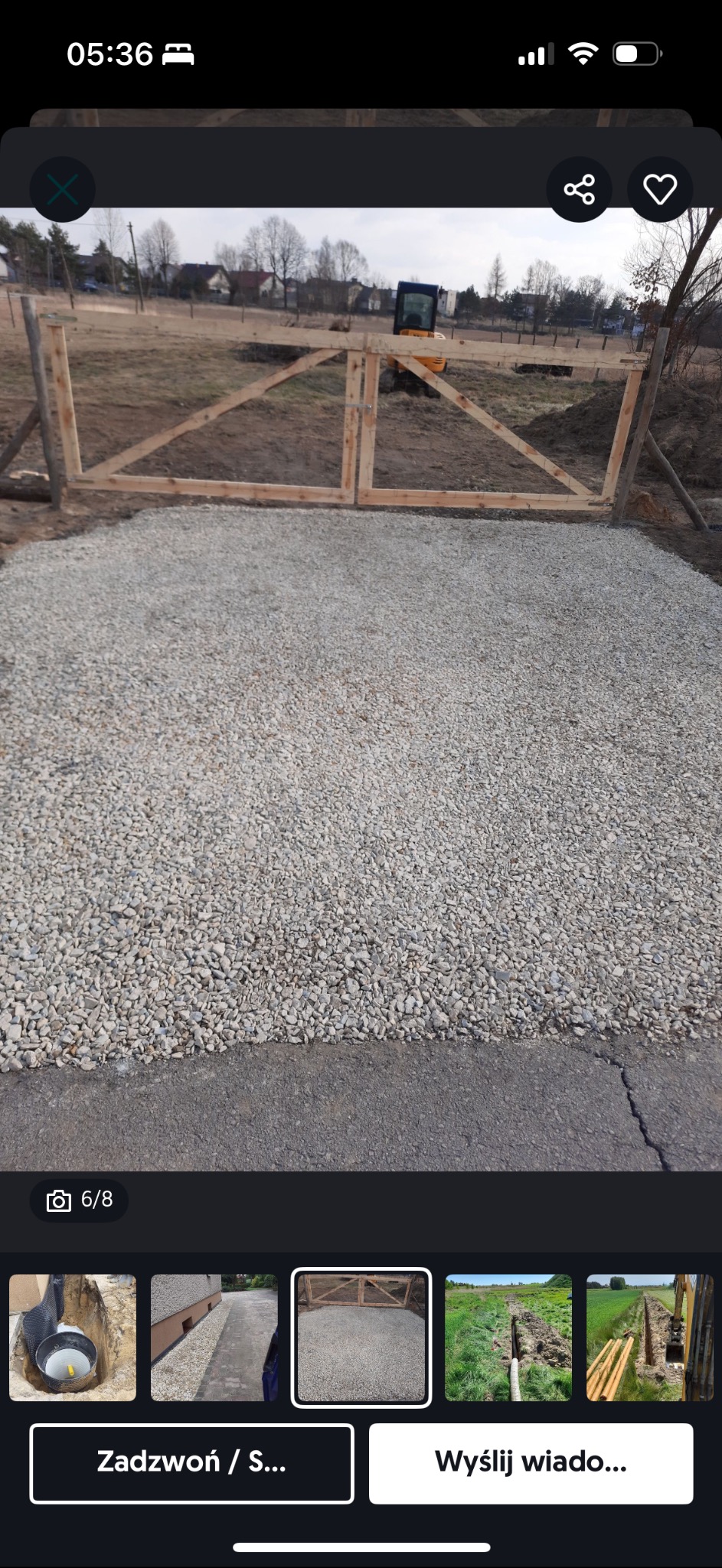 Świeżo wysypany żwir na podłożu, z drewnianą konstrukcją bramy w tle i widoczną w oddali minikoparką.