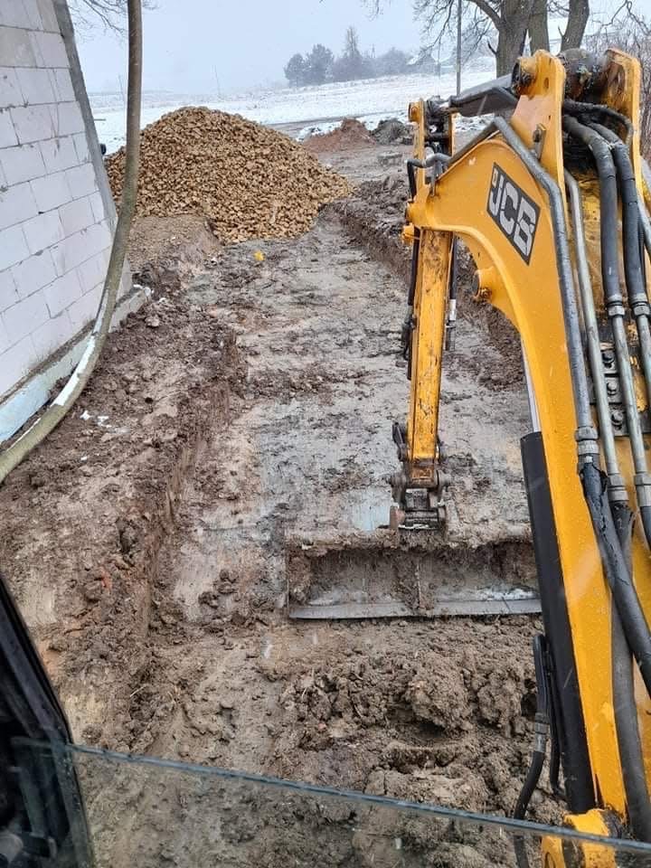 Żółta minikoparka JCB w trakcie wykonywania wykopu pod fundamenty, widoczny stos kamieni i fragment muru z bloczków betonowych, zimowa sceneria.