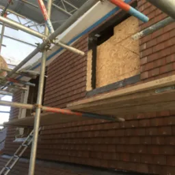 Montaż brązowej dachówki na budynku w trakcie budowy. Widoczne rusztowanie, deski i fragment izolacji. Część dachu zabezpieczona płytą OSB.