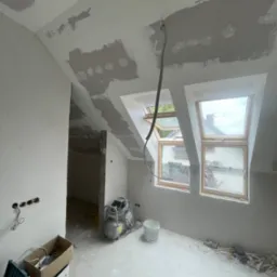 Poddasze w trakcie prac wykończeniowych: szpachlowane ściany i sufit, okna dachowe, narzędzia budowlane, widoczne przewody elektryczne. Wnętrze przygotowane do dalszych prac.