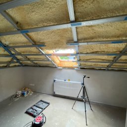 DONE AND DUSTED KONRAD SZEWCZYK - Poddasze po ociepleniu pianką PUR: widoczne metalowe profile pod płyty gipsowe, okno dachowe i narzędzia na podłodze. Etap prac wykończeniowych.