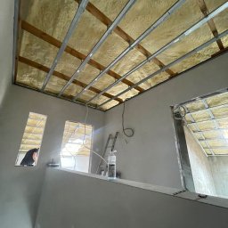 DONE AND DUSTED KONRAD SZEWCZYK - Poddasze w trakcie ocieplania pianką PUR, widoczne metalowe profile i częściowo wypełniona pianką przestrzeń. W tle drabina i osoba wyglądająca przez okno.