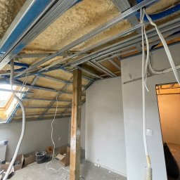 Done and Dusted - Montaż metalowej konstrukcji sufitu podwieszanego na poddaszu, ocieplonego wełną mineralną. Widoczne przewody instalacyjne i narzędzia na podłodze.