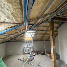DONE AND DUSTED KONRAD SZEWCZYK - Poddasze w trakcie ocieplania pianką PUR; widoczne metalowe profile konstrukcji sufitu podwieszanego i częściowo zaaplikowana pianka. Na podłodze narzędzia i materiały.