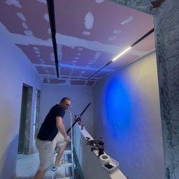 DONE AND DUSTED KONRAD SZEWCZYK - Montaż oświetlenia na suficie z płyt kartonowo-gipsowych w trakcie wykończenia wnętrza. Widoczny pracownik na drabinie i narzędzia.