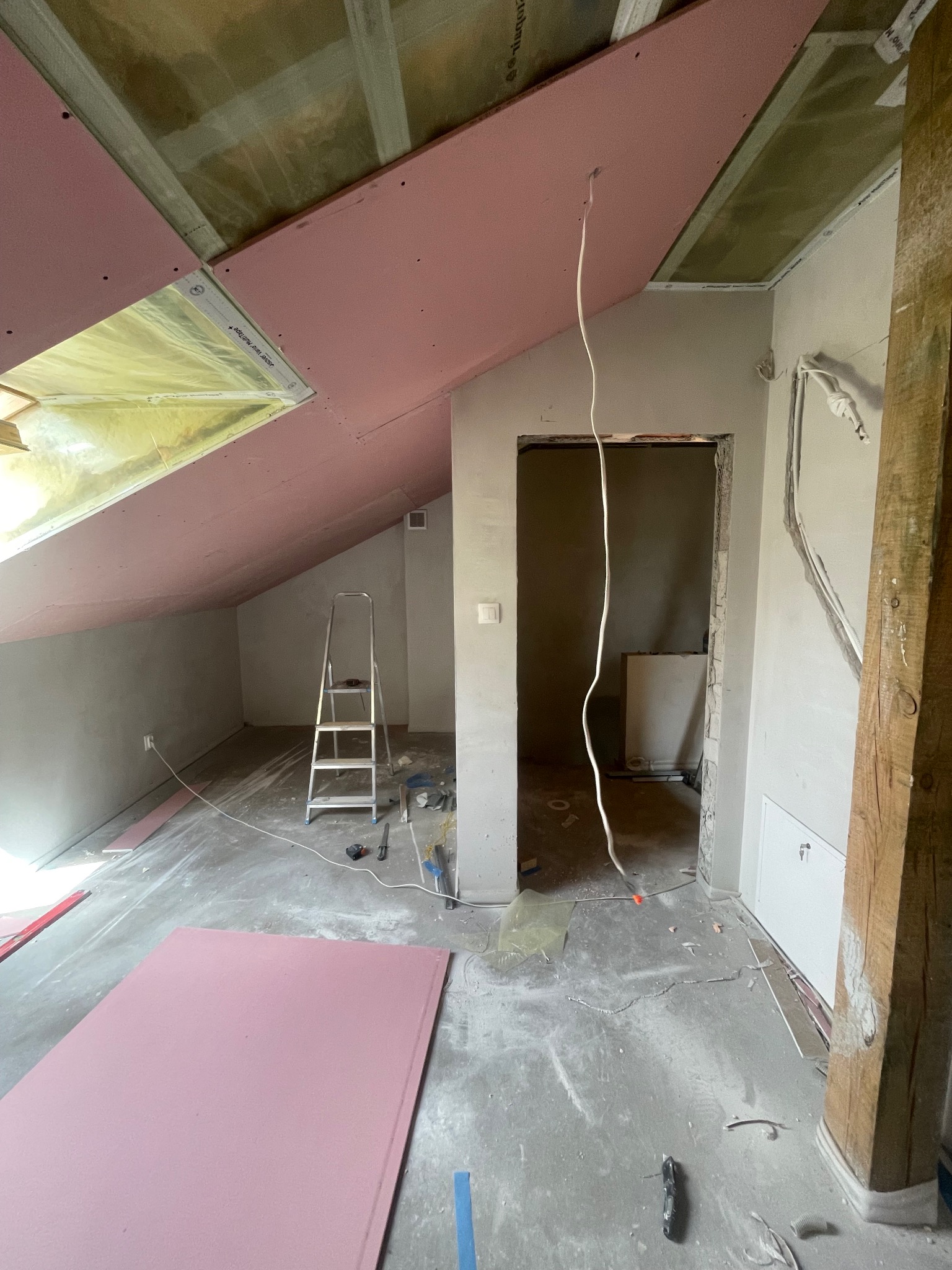 Adaptacja poddasza: montaż różowych płyt karton-gips na skośnym suficie, widoczne narzędzia i drabina, surowy stan wnętrza. Przewody elektryczne zwisają.
