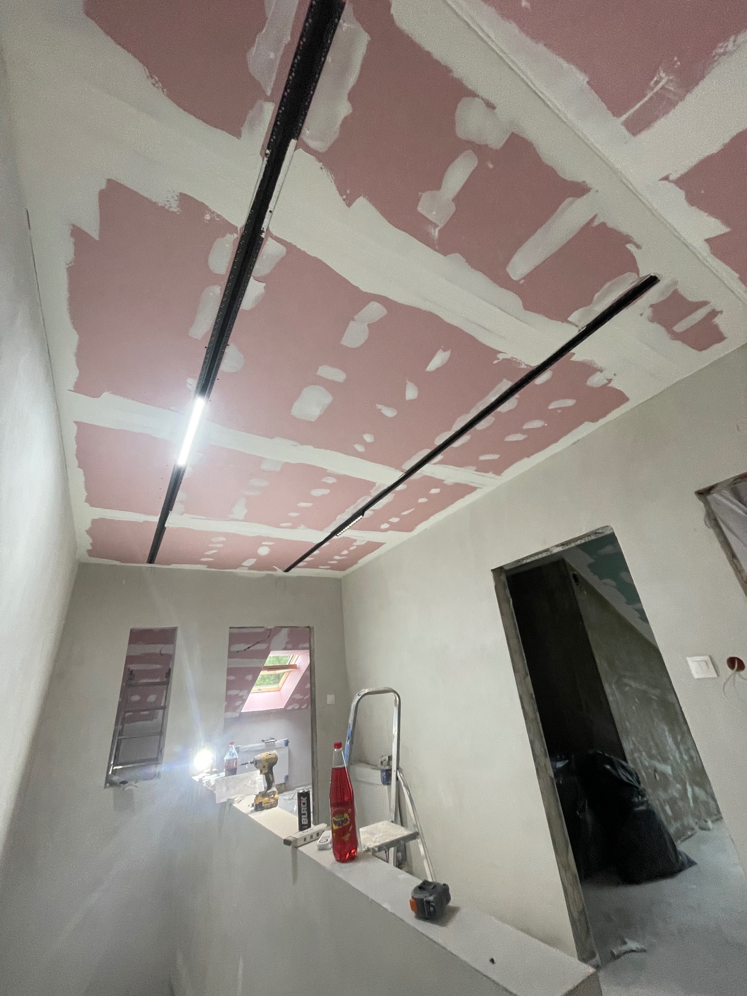 Montaż profili oświetleniowych na suficie z płyt kartonowo-gipsowych, częściowo wyszpachlowanych. Widoczne narzędzia i drabina. W tle okno dachowe i otwarte przejście.