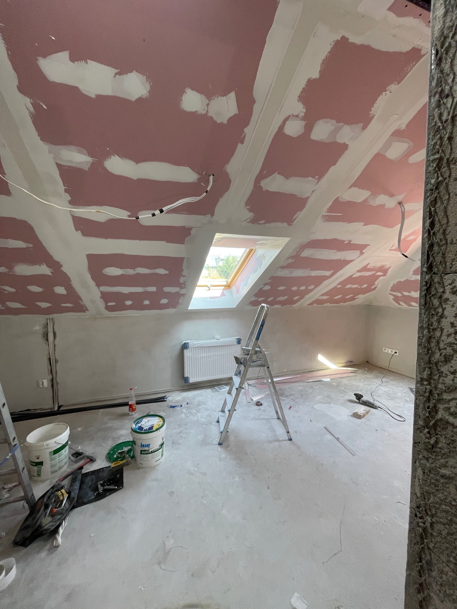 Poddasze w trakcie prac wykończeniowych: widoczne płyty karton-gips na skosach dachu, częściowo zaszpachlowane, drabina oraz narzędzia malarskie na podłodze.