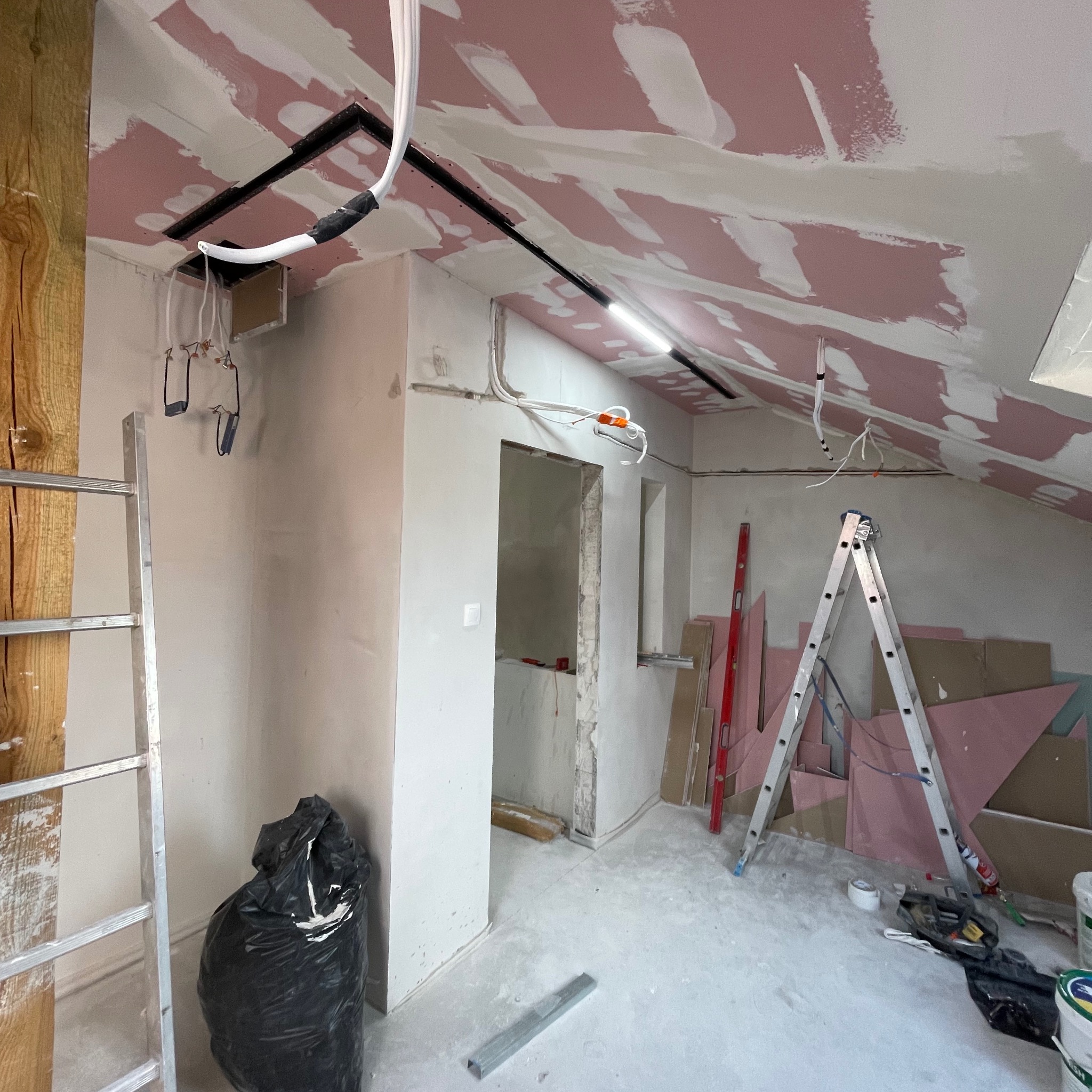 Poddasze w trakcie remontu: częściowo wykończone płyty karton-gips na skosach sufitu, widoczne instalacje elektryczne, drabina, narzędzia i materiały budowlane.