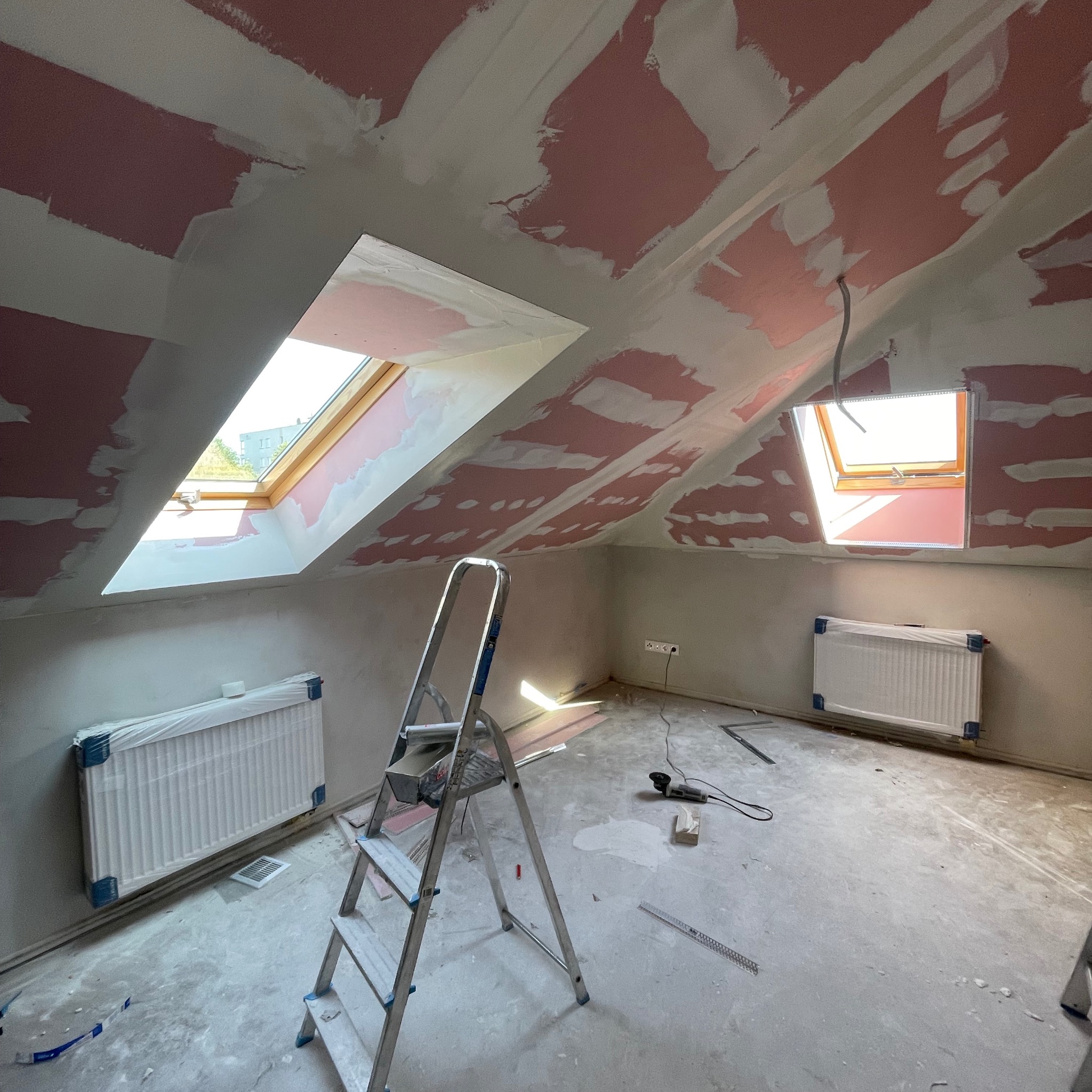 Adaptacja poddasza: sufit i ściany z płyt gipsowo-kartonowych w trakcie szpachlowania, widoczne okna dachowe, grzejniki zabezpieczone folią i drabina.