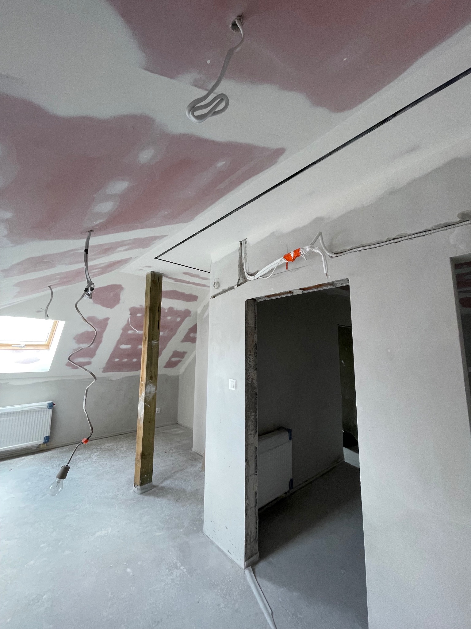 Poddasze w trakcie wykańczania: płyty gipsowe na skosach, widoczne kable elektryczne, drewniany słup podtrzymujący, surowe ściany i wylewka, okno dachowe.