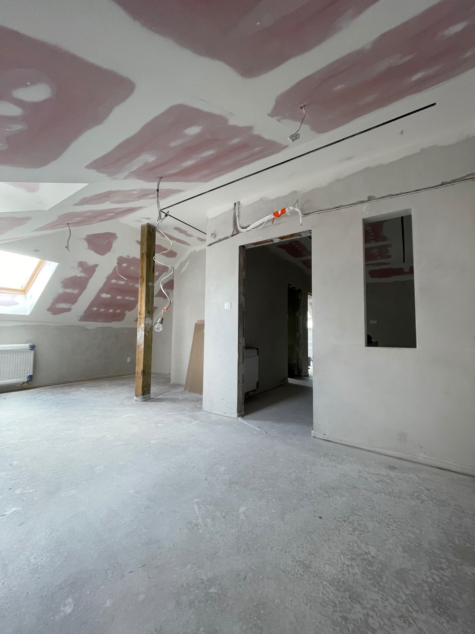 Surowe wnętrze poddasza w trakcie remontu: widoczne płyty karton-gips na skosach, drewniany słup, instalacja elektryczna i wylewka podłogowa. Okno dachowe w tle.