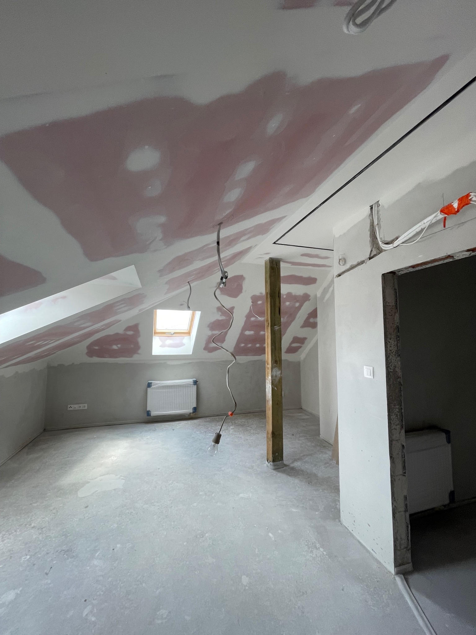 Poddasze w trakcie prac wykończeniowych: płyty karton-gips na skosach i suficie z widocznymi łączeniami, surowa podłoga, okno dachowe i drewniany słup.