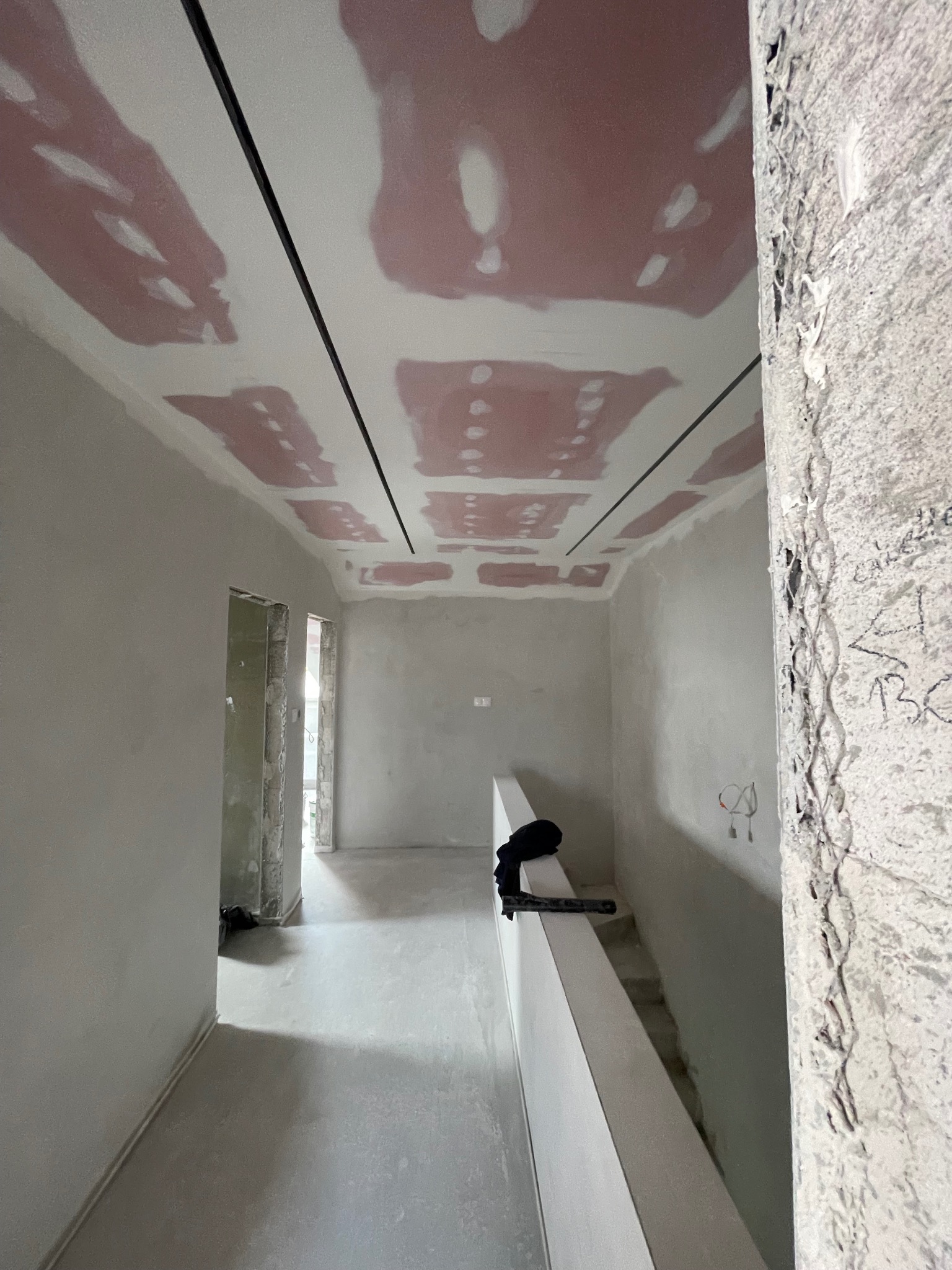 Wnętrze w trakcie prac wykończeniowych: surowy filar, gładzie na suficie z czarnymi listwami, ściany przygotowane do malowania, widok na dalsze pomieszczenia.