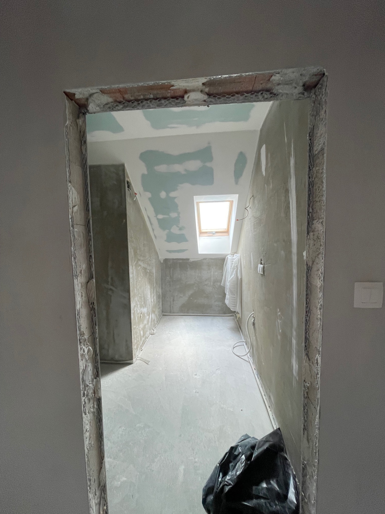 Pokój na poddaszu w trakcie remontu: surowe ściany, okno dachowe, widoczna konstrukcja drzwi. W tle widoczna instalacja elektryczna i fragment worka na śmieci.