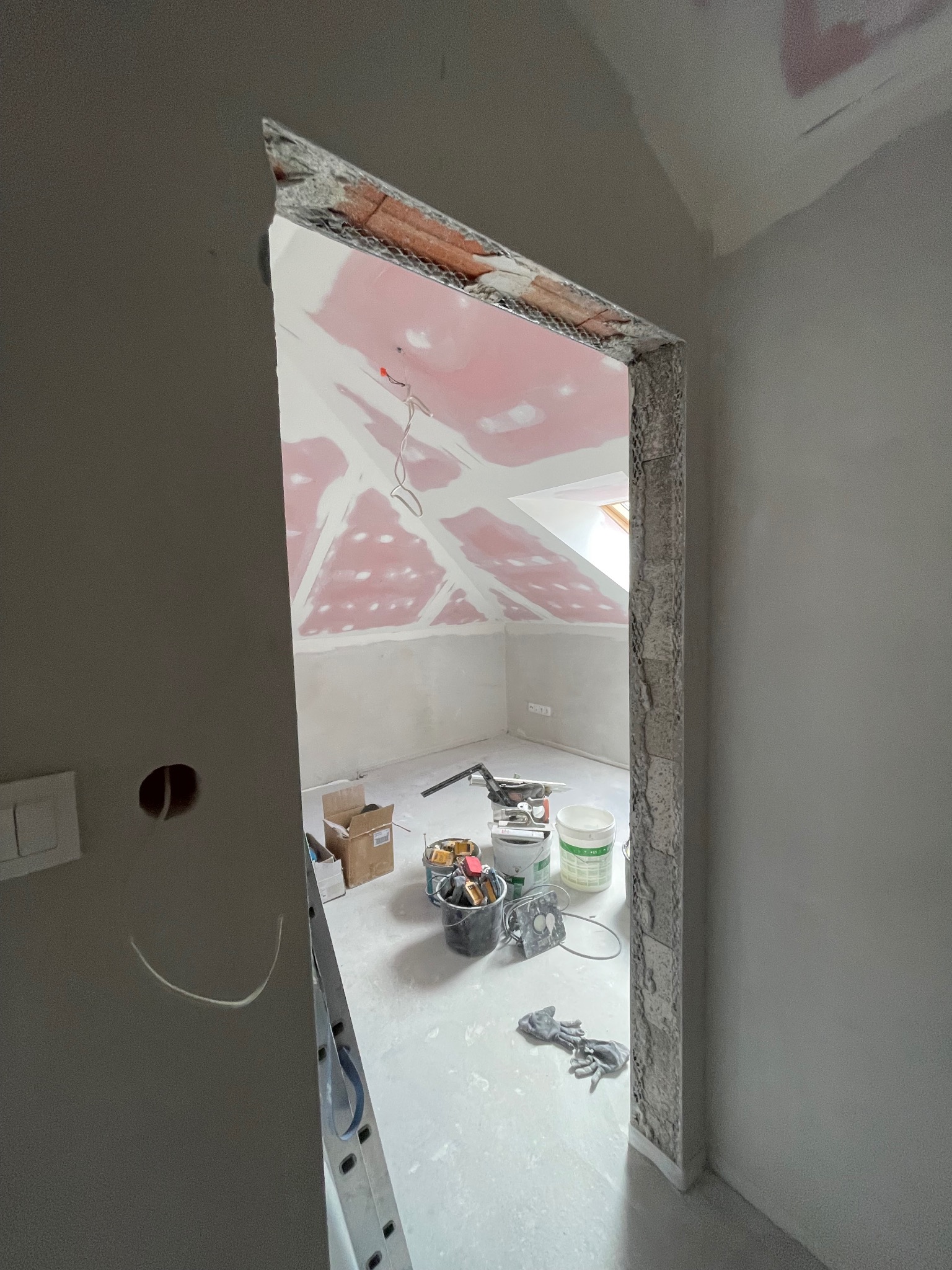 Poddasze w trakcie wykańczania: widoczne płyty karton-gips na skosach, surowe obramowanie drzwi z cegieł, narzędzia i puste wiadra na podłodze. W tle okno dachowe.