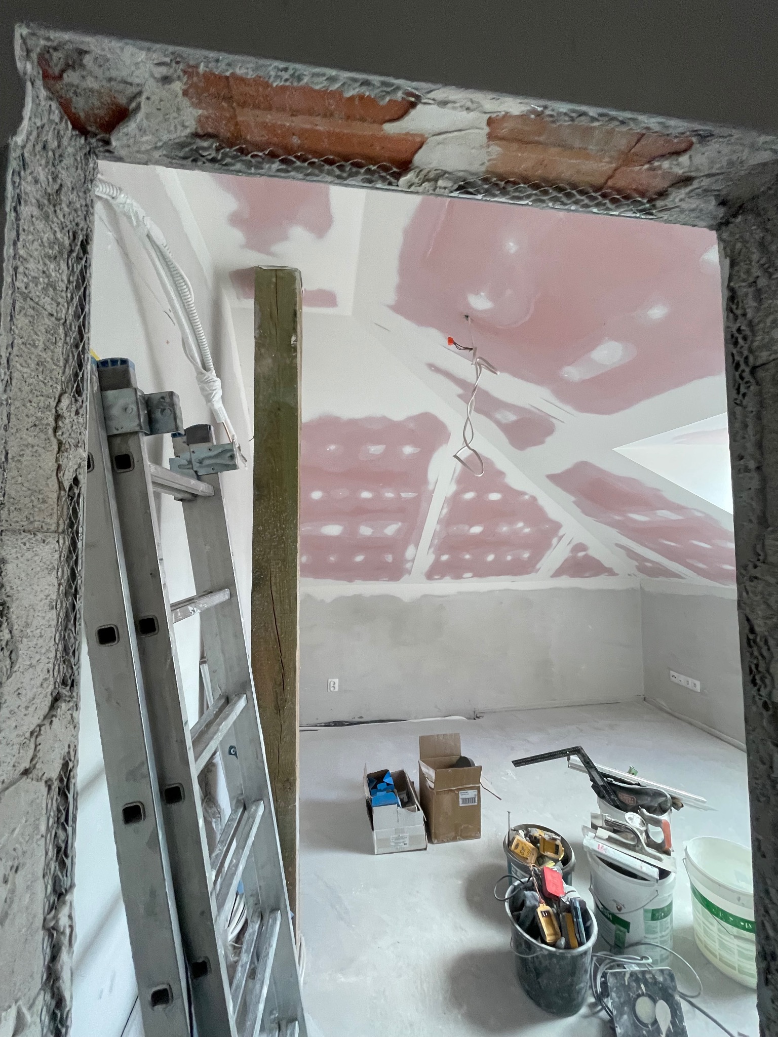 Montaż płyt G-K na poddaszu: widoczny stelaż, częściowo wykończone ściany, drabina oparta o ścianę, narzędzia w wiadrach, sufit ze skośnymi elementami.