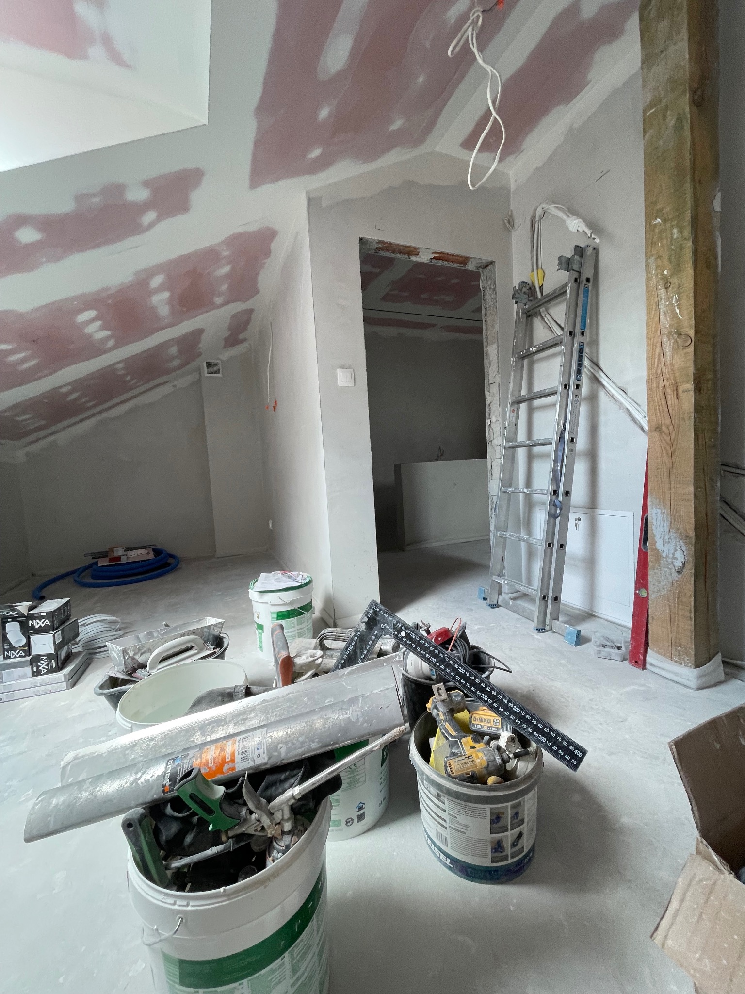 Pokój na poddaszu w trakcie prac wykończeniowych: ściany przygotowane pod malowanie, widoczne narzędzia i drabina. Skosy z płytami g-k, surowy stan.
