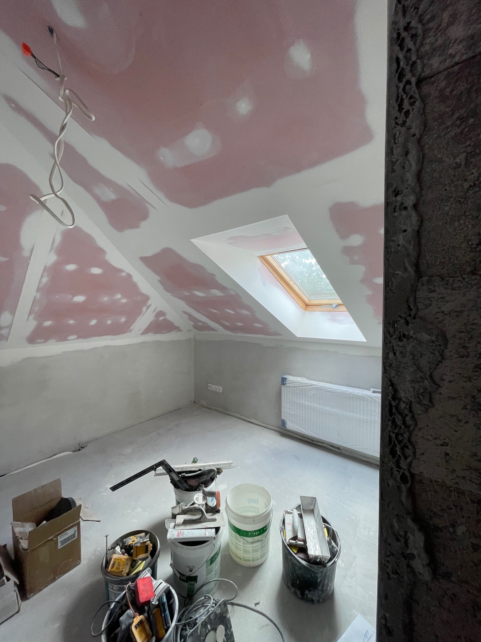 Poddasze w trakcie prac wykończeniowych: szpachlowanie płyt gips-karton, widoczne narzędzia i puszki farb. Okno dachowe wpuszcza światło.