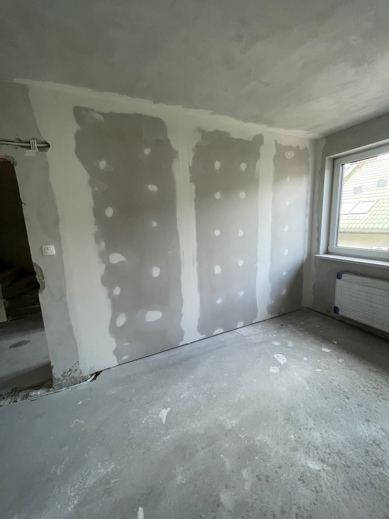 Pokój w trakcie prac wykończeniowych: ściany z płyt kartonowo-gipsowych, widoczne łączenia i miejsca po wkrętach, surowa podłoga, okno z grzejnikiem.