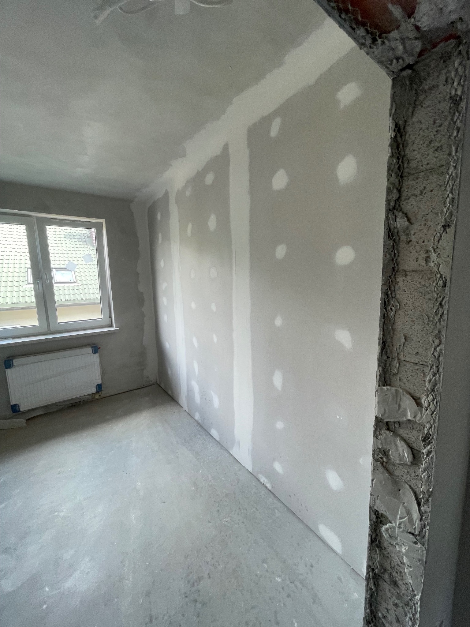 Pokój w trakcie remontu: ściany z płyt kartonowo-gipsowych, szpachlowane, surowy betonowy filar, okno z widokiem na dachy, betonowa podłoga. Etap prac wykończeniowych.
