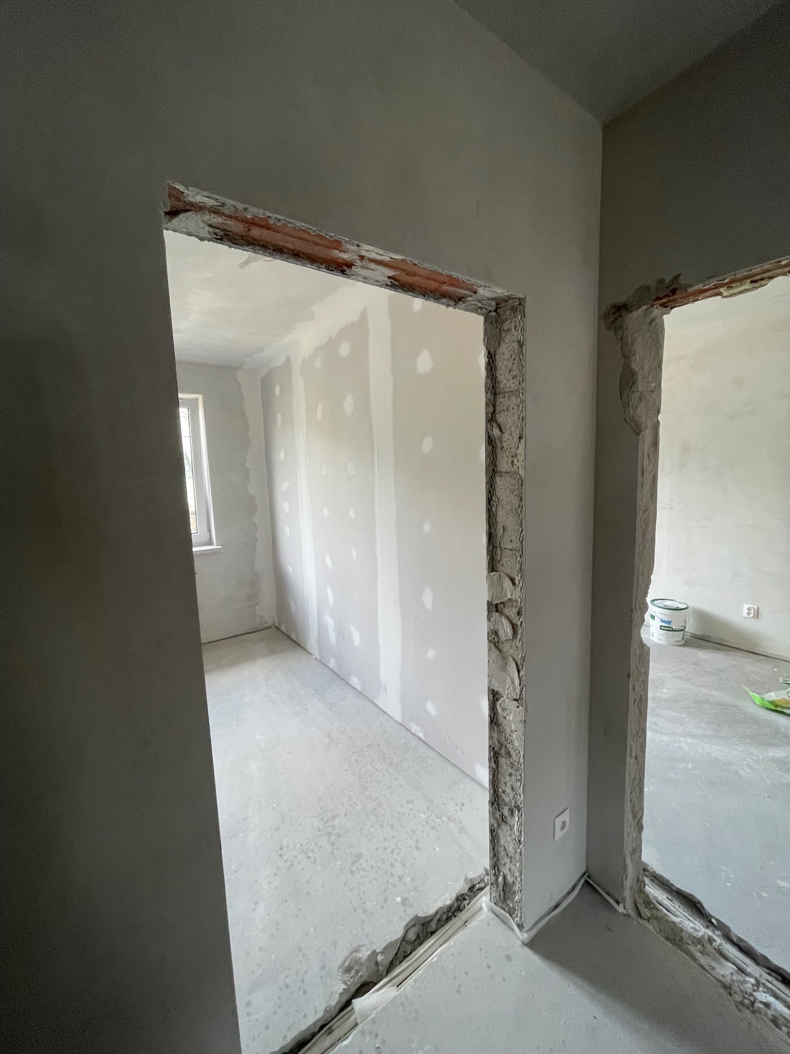 Wnętrze w trakcie remontu: ściany z płyt karton-gips, surowy beton, przygotowane otwory drzwiowe z odzyskaną cegłą. Widoczna puszka elektryczna i wiadro farby.