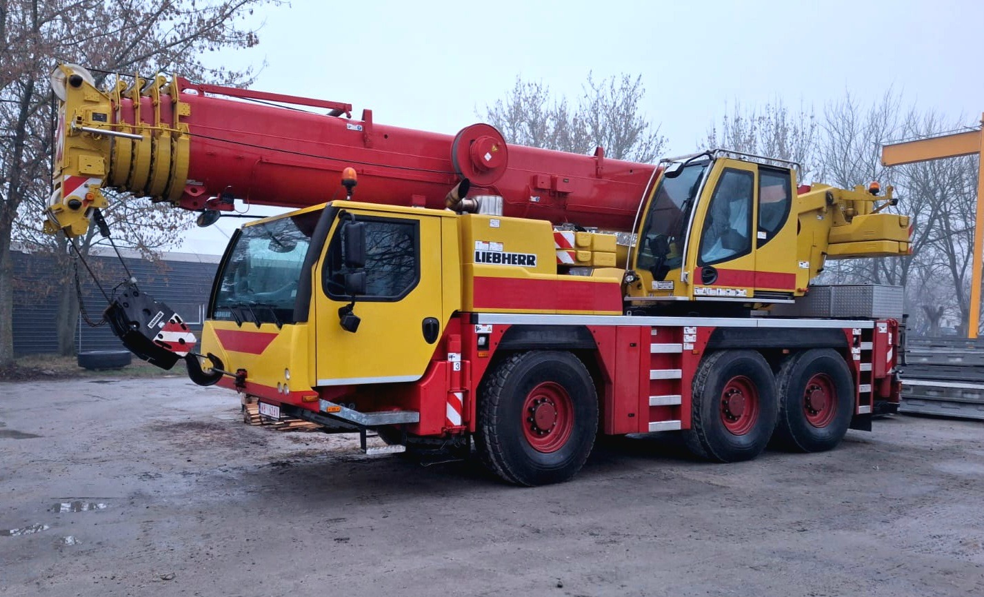 Żółto-czerwony dźwig samochodowy Liebherr na placu, widok z boku, z wysuniętym ramieniem, w tle drzewa bez liści i stalowe elementy konstrukcyjne.