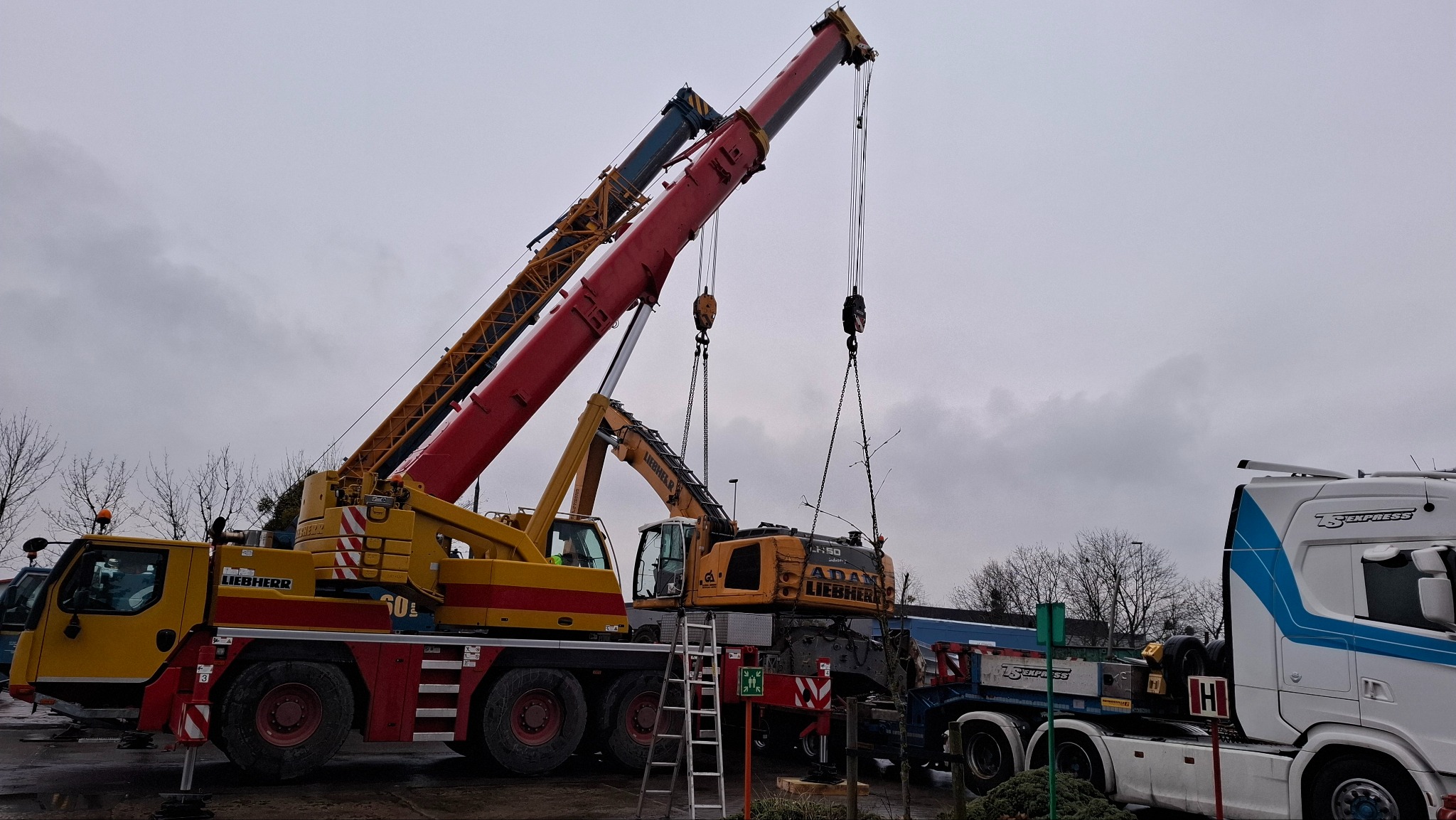 Dwa żurawie Liebherr, jeden żółto-czerwony, drugi żółty, podnoszą element konstrukcyjny na tle pochmurnego nieba, z widoczną drabiną i ciężarówką z napisem TS Express.