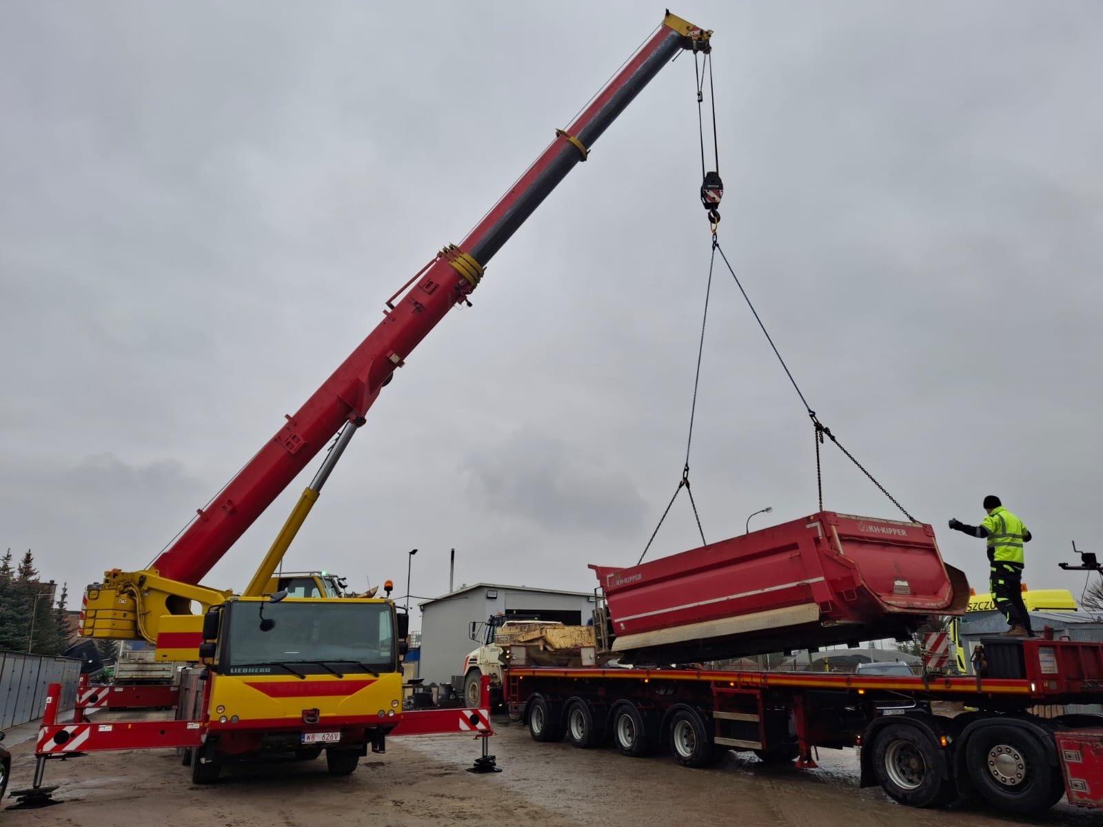 Żółto-czerwony dźwig Liebherr podnosi czerwoną skrzynię ładunkową KH-Kipper, pracownik w żółtej kamizelce nadzoruje operację na tle pochmurnego nieba.