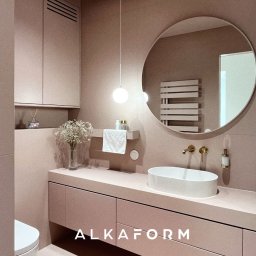 ALKAFORM - Różowa łazienka z okrągłym lustrem, białą umywalką nablatową, złotą armaturą i wiszącą lampą kulą. Minimalistyczne szafki pod umywalką i dekoracyjny bukiet gipsówki w wazonie.