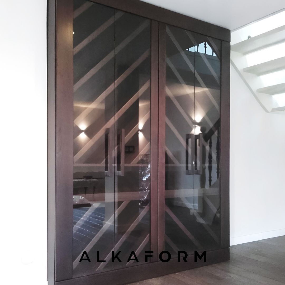 Dwudrzwiowa szafa z ciemnego drewna z geometrycznym wzorem na szklanych frontach, odbijająca wnętrze z oświetleniem i schodami.