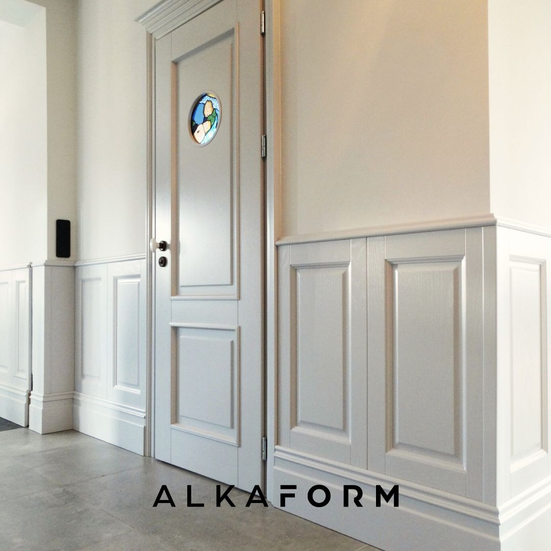 Szare, panelowe ściany z dekoracyjną stolarką, drzwi z okrągłym witrażem i listwy przypodłogowe w jasnym wnętrzu.