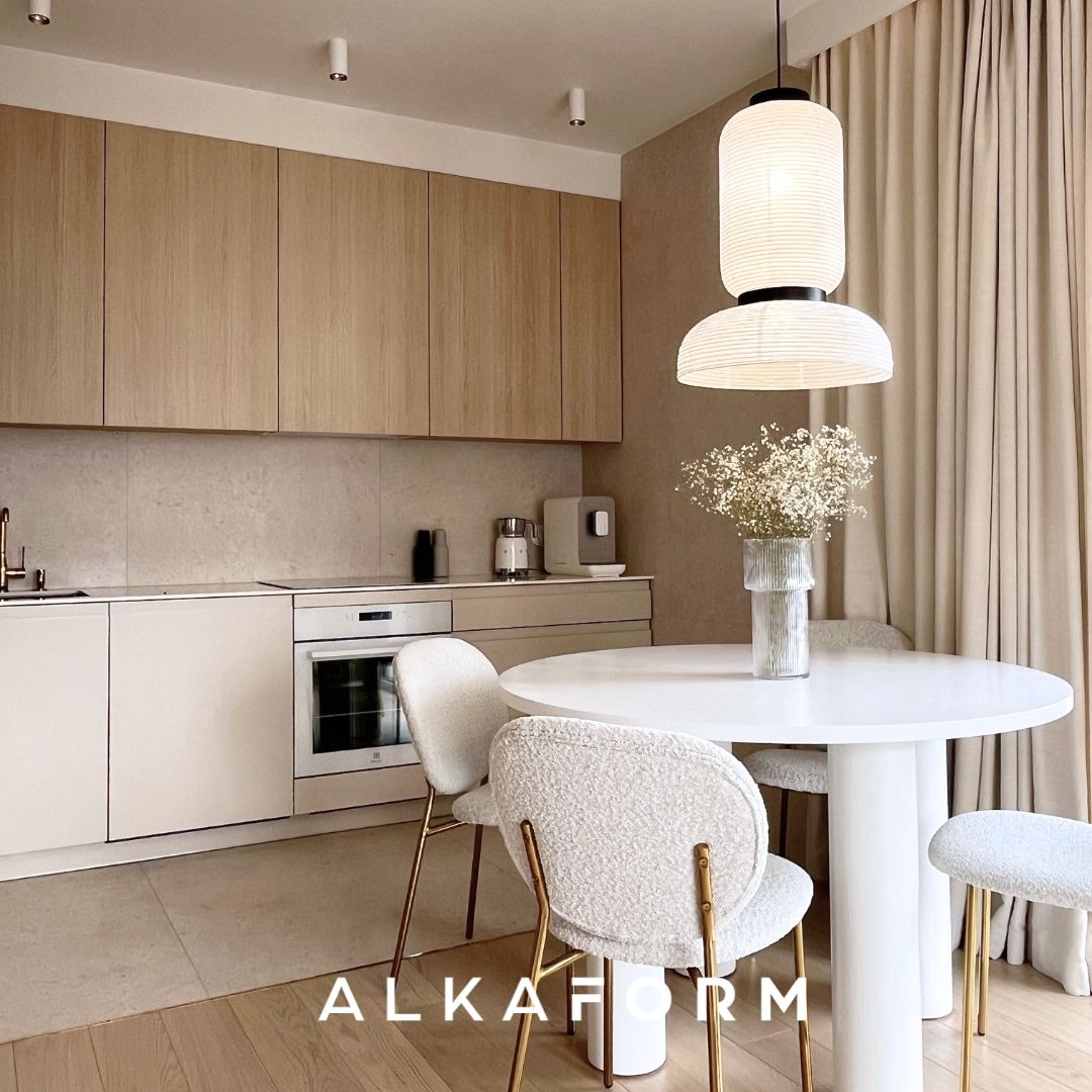 Jasna kuchnia z drewnianymi szafkami, białymi frontami i okrągłym stołem z białym blatem i krzesłami na złotych nogach, oświetlona wiszącą lampą w stylu japońskim.