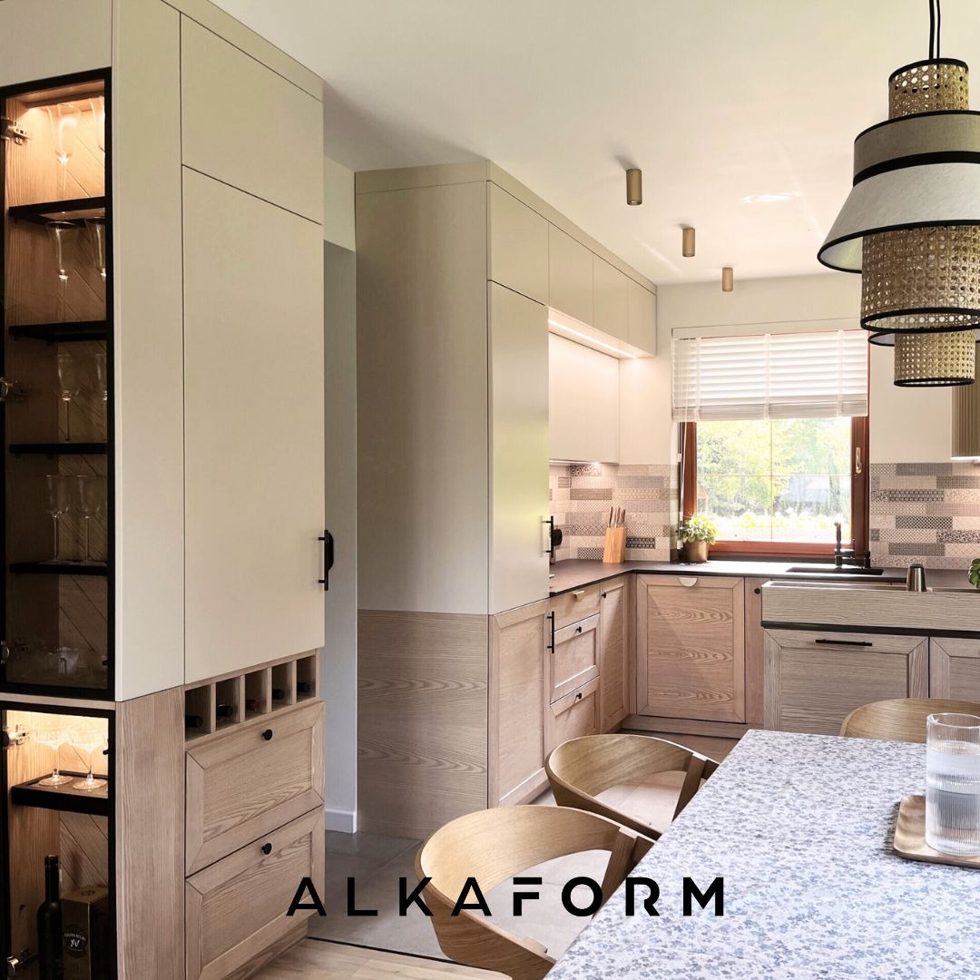 Jasna kuchnia z drewnianymi akcentami: szafki z przeszklonymi witrynami, wysuwane szuflady na wino, blat z szarego lastryko, okno z widokiem na zieleń, nowoczesna lampa wisząca i logo firmy...