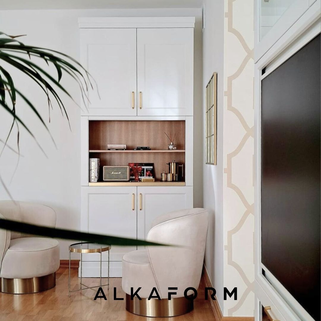 Biała zabudowa meblowa z otwartą półką, uzupełniona beżowymi fotelami na złotych podstawach i stolikiem kawowym w salonie z drewnianą podłogą.