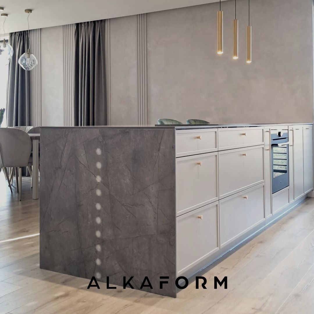 Minimalistyczna wyspa kuchenna z szarym blatem imitującym kamień i białymi szafkami z delikatnymi złotymi uchwytami, oświetlona trzema wiszącymi lampami w kształcie złotych tub, w tle zasłony...