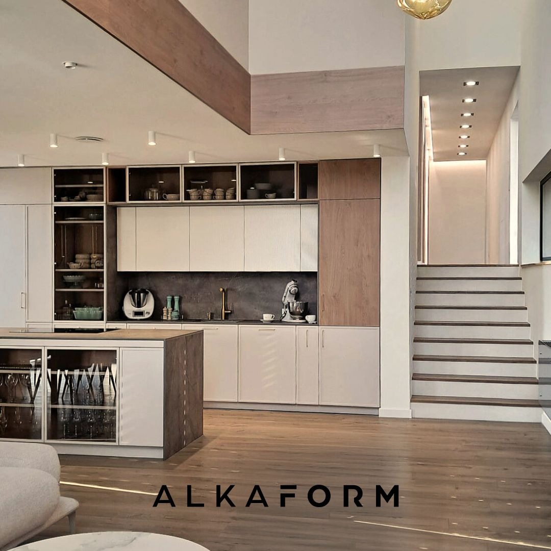 Nowoczesna kuchnia z wyspą, jasne fronty szafek, drewniane akcenty, przeszklona witryna na wyspie kuchennej, schody w tle, oświetlenie punktowe, logo firmy ALKAFORM na pierwszym planie.