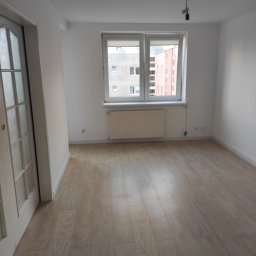 Budowa - Pusty pokój z nową podłogą z paneli laminowanych, białe ściany, okno z widokiem na budynki, białe drzwi z przeszkleniami po lewej stronie.