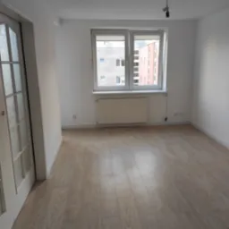 Pusty pokój z nową podłogą z paneli laminowanych, białe ściany, okno z widokiem na budynki, białe drzwi z przeszkleniami po lewej stronie.