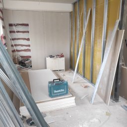 Budowa - Montaż ścian działowych z płyt karton gips z widoczną izolacją, profile stalowe, narzędzia Makita i stos płyt gipsowych w surowym wnętrzu.