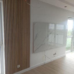 Budowa - Ściana z drewnianych paneli akustycznych, imitacja marmuru na ścianie, widoczne kable i gniazdka elektryczne, podłoga z paneli.