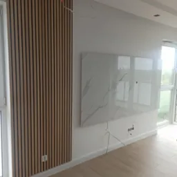 Ściana z drewnianych paneli akustycznych, imitacja marmuru na ścianie, widoczne kable i gniazdka elektryczne, podłoga z paneli.