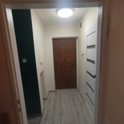 Budowa - Korytarz po remoncie: szare panele podłogowe, białe ściany z fragmentem pomalowanym na ciemny zielony kolor, brązowe drzwi wejściowe i białe drzwi wewnętrzne z poziomymi wgłębieniami.