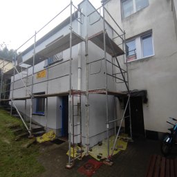 Budowa - Budynek mieszkalny w trakcie odświeżania elewacji, z rozstawionymi rusztowaniami, zabezpieczonymi oknami i żółtą folią ochronną na ziemi.