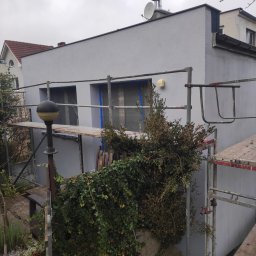 Budowa - Świeżo pomalowany, szary budynek z rusztowaniem i zabezpieczonymi oknami, widok z zarośniętego ogrodu w pochmurny dzień.