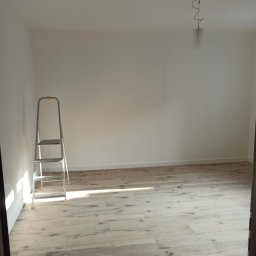 Budowa - Wnętrze pokoju po remoncie: świeżo pomalowane białe ściany, ułożone jasne panele podłogowe imitujące drewno, aluminiowa drabina oparta o ścianę, widoczny fragment sufitu z lampą.