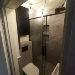 Nowoczesna łazienka z kabiną prysznicową typu walk-in, wiszącą toaletą i umywalką nablatową, wykończona płytkami imitującymi kamień, oświetlona lampami wiszącymi z żarówkami Edison.