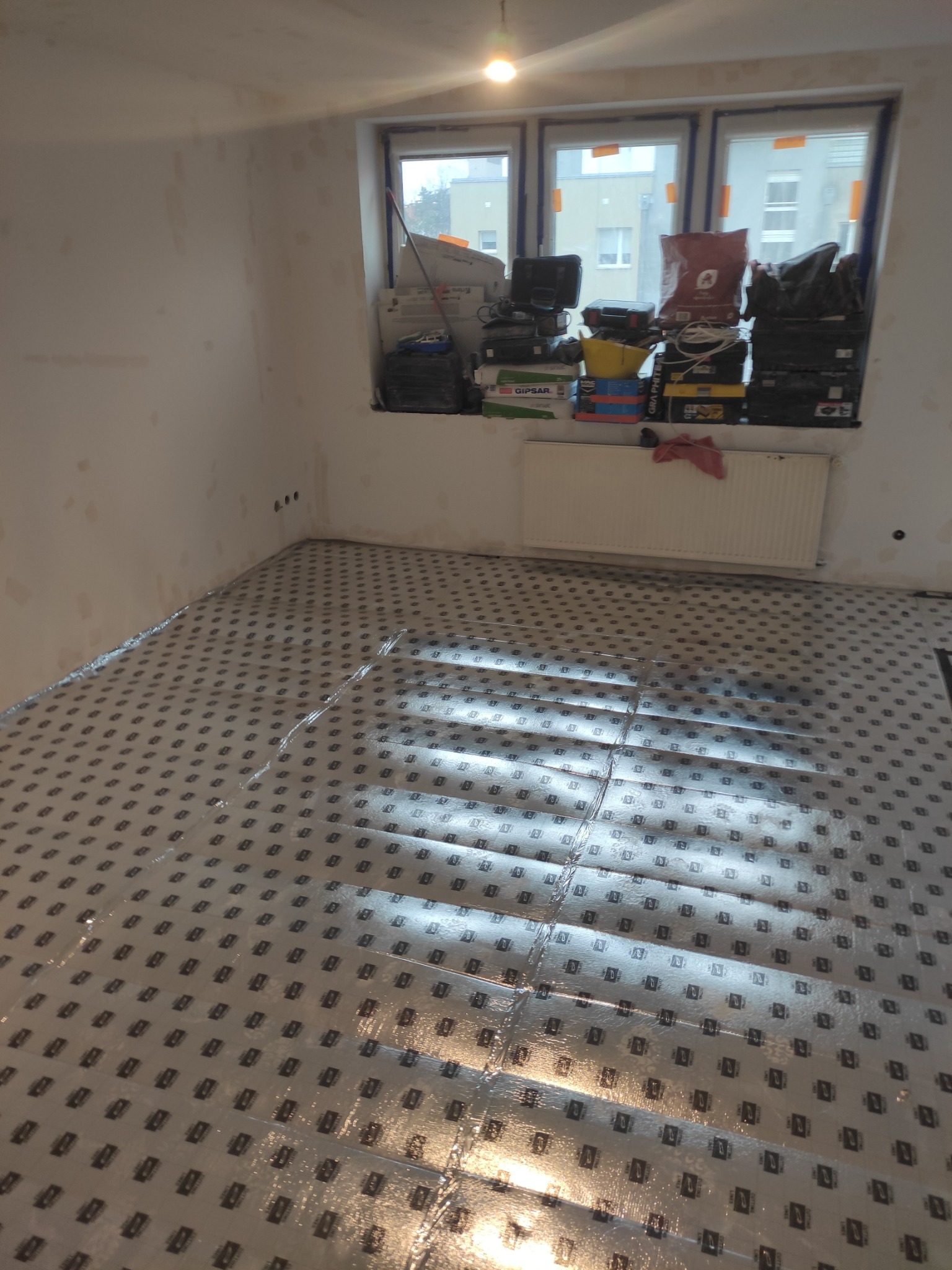 Przygotowanie podłoża pod panele: folia paroizolacyjna rozłożona na podłodze w pokoju z oknem, na parapecie narzędzia i materiały budowlane.