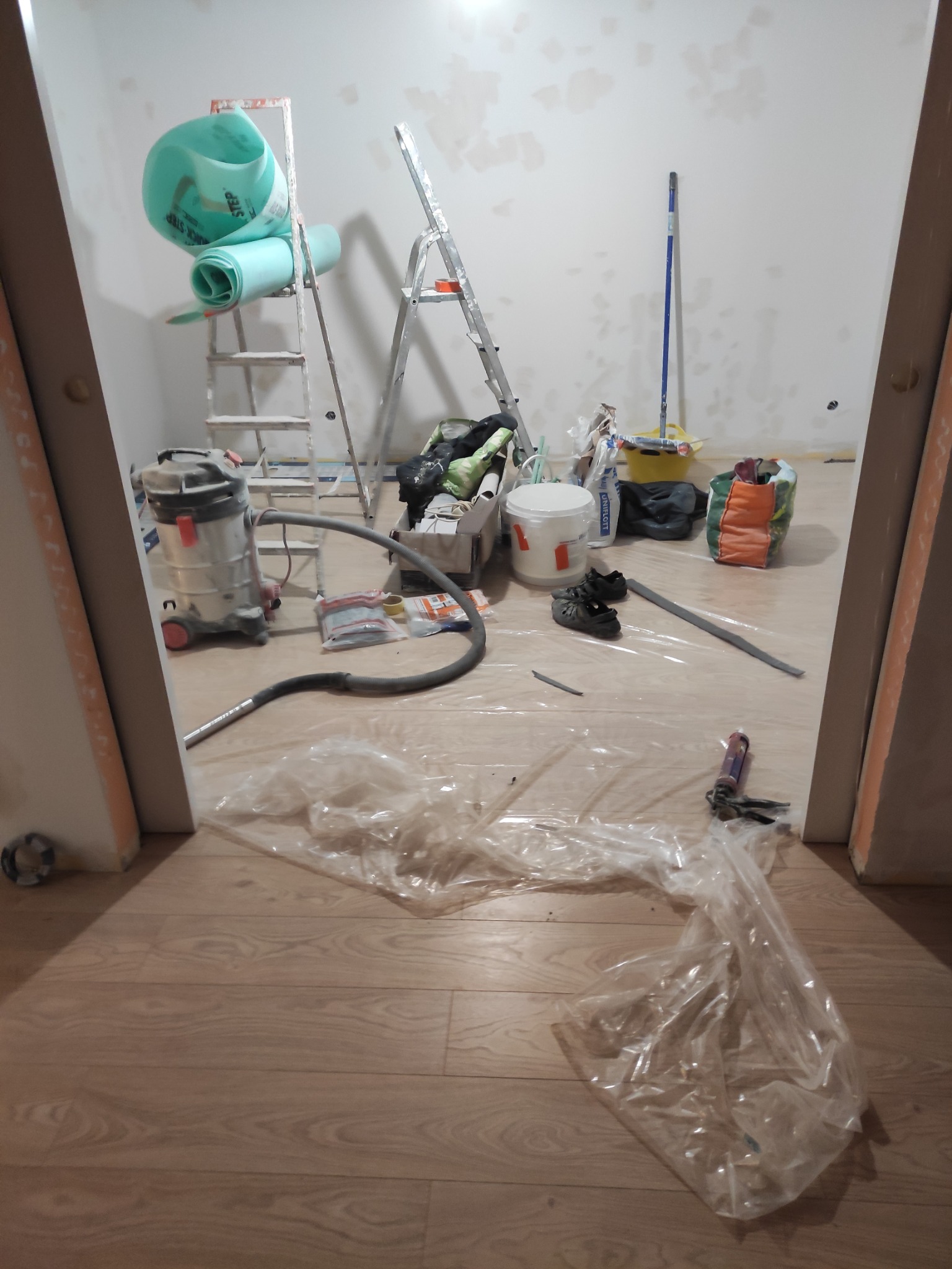 Remontowany pokój w Gdańsku z rozłożoną folią ochronną na podłodze, widoczne drabiny, rolka materiału izolacyjnego, odkurzacz przemysłowy i narzędzia.