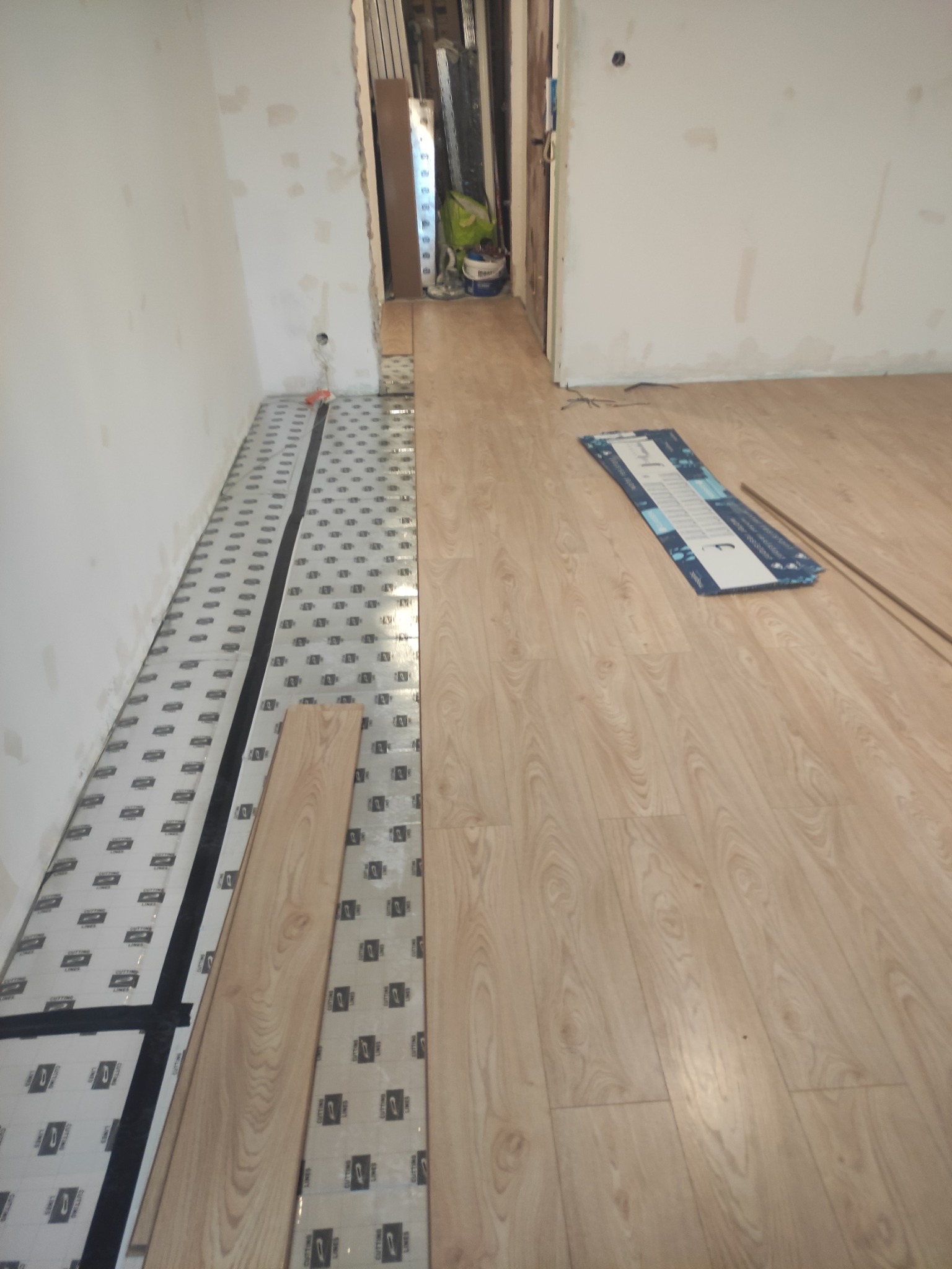 Układanie paneli podłogowych w pomieszczeniu: widoczny podkład z folią, częściowo ułożone panele o jasnym odcieniu drewna i listwa pomiarowa na podłodze.